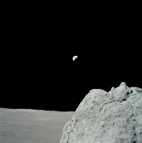 The half Earth is seen from the surface of the Moon against the deep black of space. A large boulder sits in the foreground of the photograph.