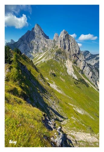 Photo of a picturesque mountain scene. A steep, grassy ridge is leading from the foreground toward a mountain pass and the vertical rocky cliffs and peaks of Kellenspitze (2238m) and Kellenschrofen (2091m) in the background. From the pass, another path is leading down the steep grassy slopes into the valley on the right. Bright sunny, blue sky and some fluffy clouds moving in and casting shadows on the peaks.
