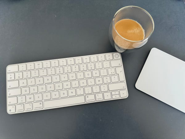 Sur un bureau noir, sur la gauche, un clavier, Apple, blanc portant des inscriptions en noir, sur la droite, un trackpad Apple blanc, et entre les deux, une tasse de café brun mais noirdans une tasse en verre.