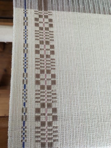 close up on the edge of handwoven cloth on a loom. the ground cloth is grey and taupe, with brown monksbelt motifs along the selvedge. the outer motif is narrow, with a thin blue stripe in the center. the inner motif is a traditional monksbelt rose pattern with a thin pink stripe in the center.