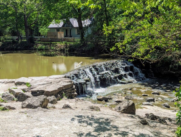 Photo of a small waterfall spanning a wide stretch of a small creek. An old house or mill building sits on the other side of the creek 