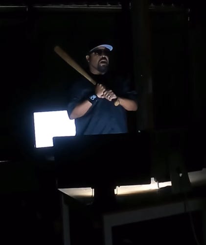 Photo. A color photo of the black rapper "Ice Cube". The man has a full beard, wears sunglasses and a black baseball cap and is dressed in black.  He is standing in front of some computer screens in the dark, holding a baseball bat in his hand. A little light falls on his face. He is about to take a swing and smash the computer.
Info: There is a mural by the artist Royyal Dog of this action by the two artists.