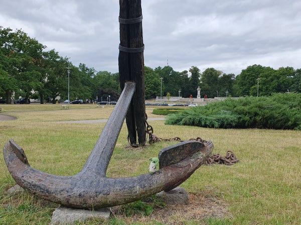 A large, used, rusting anchor sits on the grass in a park, trees and a monument in the background. Grogu leans back against the anchor.