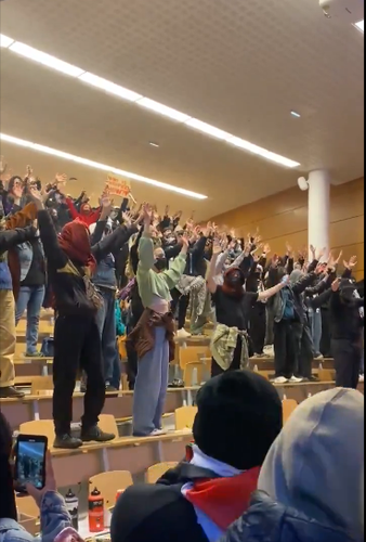 A large number of students standing on their desks with their arms in the air