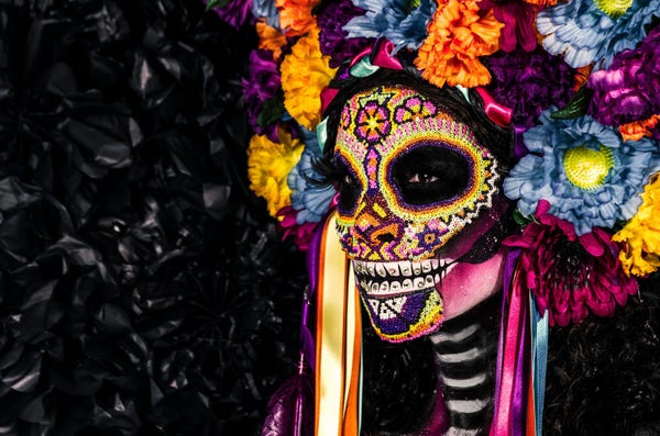 Unsplash photo by fer gomez of a woman dressed in an elaborate Dia de los Muertos costume: a colorful painted skull face with a headdress of flowers and ribbons. 
