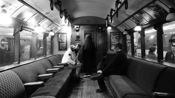 black and white interior of a 1930s tube carriage. Two young men are sat in opposite seats talking and smiling. Beyond them at the end of the carriage a mysterious girl with long dark hair is facing the carriage end window. Her body is in shadow.