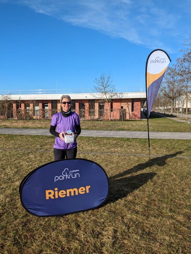 Yours truly wearing purple tshirt for International Women's Day, holding a box of chocolate cakes I baked. I stand behind the Riemer pop-up sign. The Parkrun flag is on the right hand side