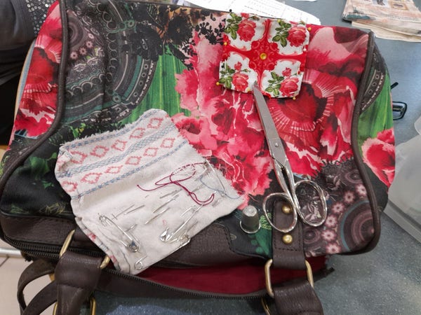 A handbag with a strong-coloured and abstract design of roses in green, red, and black. On top is a patch with a geometric rose pattern, a collection of sewing needles and a pair of scissors.
