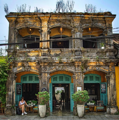 Charming old Vietnamese building, painted gold with a combination of french architecture and vietnamese. The building is delapidated and covered in mold. The building has been somewhat restored, green shutters, comfy chairs and potted plants. very inviting scene.