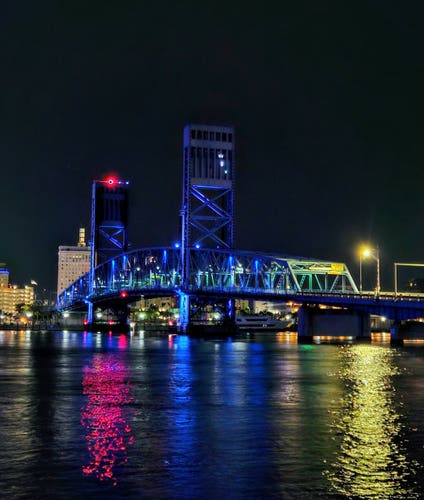 Late night view across the Saint Johns River at the main street bridge,  illuminated in blue and reflecting with colorful traffic lighting upon the calm dark waters below.