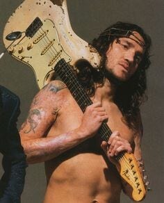 John Frusciante torse nu, portant sa guitare par le manche sur le dos, comme un montant de croix
