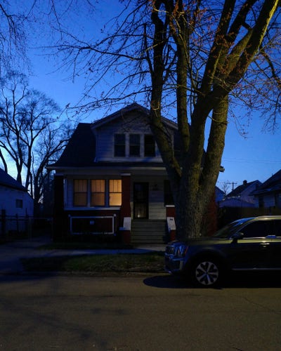 Color photograph looking across a street at dusk at the front of a wooden house, a row of three windows glowing with yellow light with the rest of the face in shadow. A large tree with several large thick branches stands in front of the house, light cast against it from the right