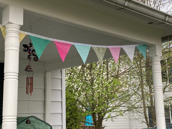 A string of colorful bunting flags hung on a porch, with a wind chime, tree blossoms in the background, and a corner of a house visible.