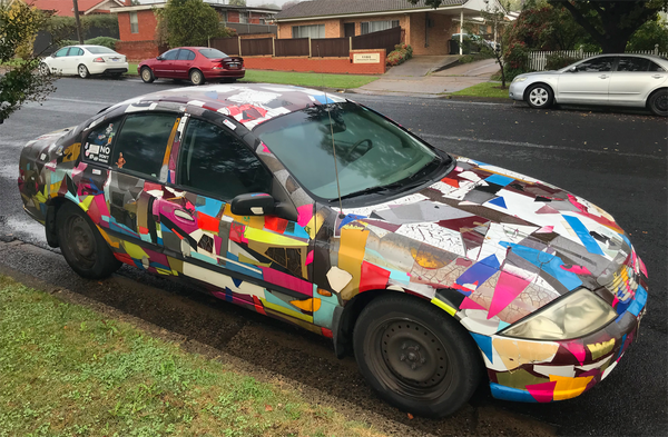 A photo of a car parked in a street absolutely covered in roughly cut pieces of colourful signage vinyl. One end to the other, it's all kinds of colours, and different kinds of vinyl. Some have aged with a fine patina, others almost falling off, others still in good condition. It's simply an art car.