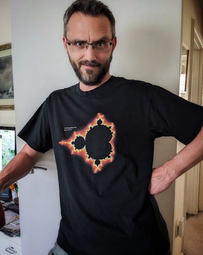 White male, dark short hair and beard, wearing a black t-shirt with a print featuring the Mandelbrot set fractal
