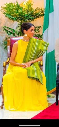 Meghan Markle sits in a bright yellow dress with a colourful lime green striped green sash. She sits in front of the Nigerian flag.