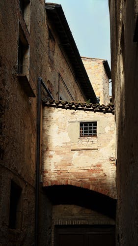 A brick passageway connects two sides of a narrow street in Pienza. A patch of sunlight illuminates its solitary small, square window which is protected by a grid of iron bars.