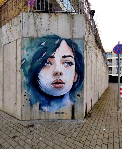 Streetartwall. A charming little portrait of a young woman has been painted with acrylic paint on a desolate corner concrete wall. She has wavy black hair, a pale face, full lips and is gazing longingly into the distance. The background is blurred blue . (In the photo you can see a sidewalk of cobblestones in front of it, a rusty balcony grille above it and a traffic sign on the right-hand side).