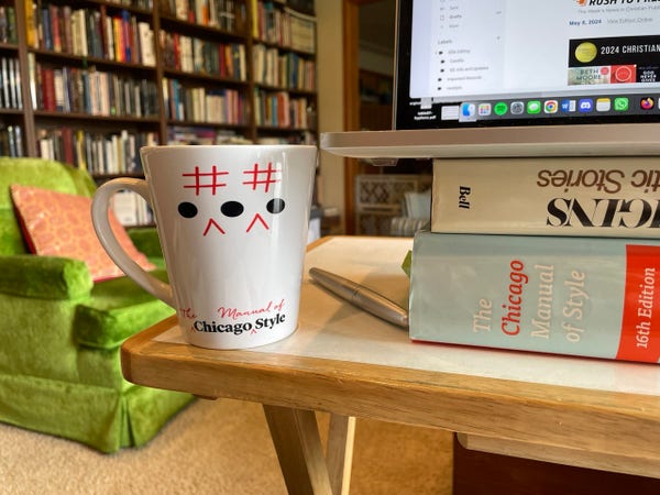 A mug sporting big, black ellipses sits on a TV tray next to an open laptop. The ellipses have editor’s marks requesting spaces between each dot.