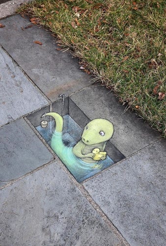 Streetart. A hole with a green dinosaur has been drawn with chalk on a rectangular slab of stone on a sidewalk. The cute dinosaur sits in the painted water with a yellow squeaky duck and takes a bath. Unfortunately, he has a long tail and the bath plug has got tangled up in it. He looks a little confused at his tail (A piece of meadow can be seen on the right of the photo.) Title: "Yet another bath night cut short by Brian's free-spirited tailbone."
