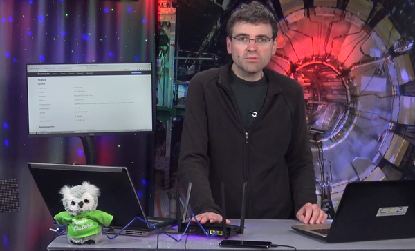 Still aus dem Video: Moderator hinter Tisch mit zwei Laptops, in der Mitte der Router, links vorne die Eule, das Maskottchen der Sendung mit einem T-Shirt, auf dem Frohe Ostern steht 