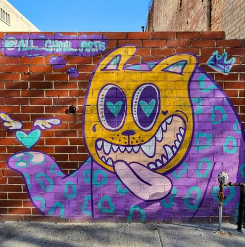 A red brick wall with a bright art/graffiti image of a cartoon animal with a yellow head, open mouth sticking its tongue out, large purple eyes with blue heart centers, and a large purple and blue body, with an arm extended to hold up a white-winged blue heart.