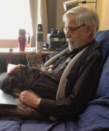 A young tabby cat is lying happily on the lap of a man.  The man has white hair, a beard and glasses.  The cat's little white mittens are resting on the man's chest.