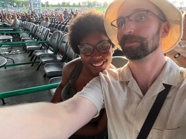 A photo of me and my wife. We are sitting in amphitheater seating, and there are empty seats beyond us. i’m wearing a tan shirt-leave button down and matching bucket hat, and she is wearing a dark green dress. 
