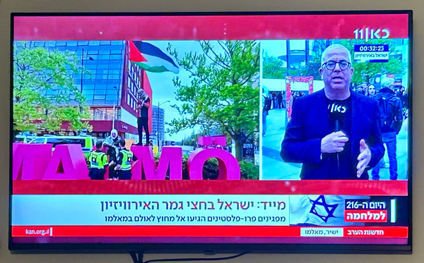 Fernseher mit Livebild von Kan|11 mit einem Moderator und einem eingeblendeten Video von einem Verhüllten Mann mit Palästina-Fahne, der auf ein Monument aus den Buchstaben MALMÖ geklettert ist.