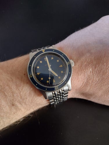 Modern 1960's style diving watch with a steel beads of rice bracelet. The watch face is minimal with little triangles pointing inwards at the 3, 6 and 9 positions; the other hours are spots except for a numerical 12. The face is a sunburst blue that reflects a different hue depending on the light, like you would find on a Fender guitar, while the hands and details are in a goldish patina