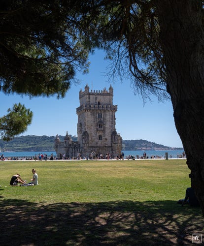 Color photo of a Portuguese Manueline-architectural-styled stone tower sitting just off the banks of a wide river mouth under blue, sunny skies. The tower is framed by a large tree and its overhanging branches and a large grassy park lawn in the foreground. 