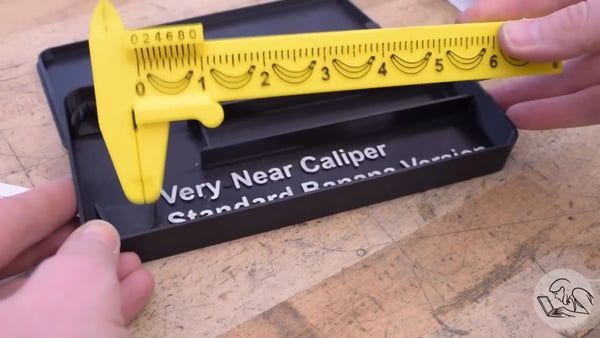 Two white hands holding a small vernier caliper made from yellow plastic. Its scale carries several pictures of bananas between the numbers. The black case below it on the workbench is obviously 3d-printed, and marked with white text saying "Very Near Caliper Standard Banana Version"