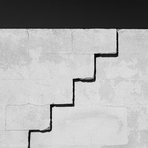 In this photograph titled "Steps," you'll find a grayscale image of a white concrete wall. The wall is marked by a prominent black crack that runs through the plaster, forming a pattern resembling stairs. It's a minimalist composition that captures the contrast between light and dark, drawing attention to the linear form of the crack against the smooth surface of the wall.