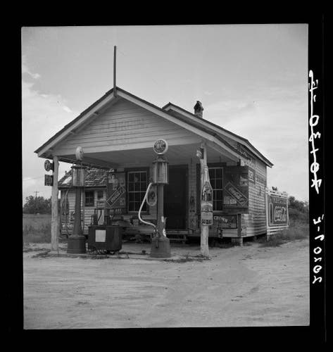  The image is a vintage photograph of a small rural gas station. It's located in Granville County, North Carolina, and features an old-fashioned structure with a shingled roof and white siding. Two gas pumps are visible on the left side of the building. There's also a sign that reads "SUNOCO" above one of the pumps. A small porch is present in front of the station, suggesting it may have served as a gathering spot for farmers in their off times. The overall scene suggests an era when gas stations were often independently owned and operated.