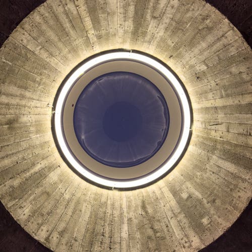 Looking up through a round skylight. It’s framed by Beton Brut and there is a lamp around the window.