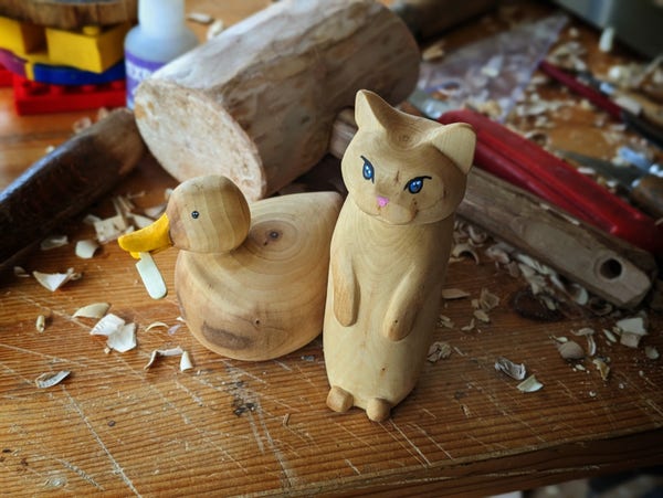 Woodcarved sculpture on a messy workbench. A duck with a buttet knife in its beak, and a standing blue eyed cat.