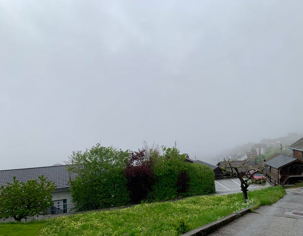 View of a street, green grass and trees but the rest of the village is covered in thick clouds and it's raining.