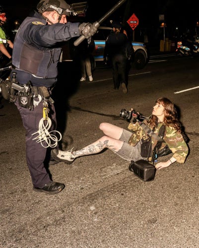 NYPD officer wielding baton stands over credentialed press Olga Federova who is lying on the ground in front of him holding her camera looking up