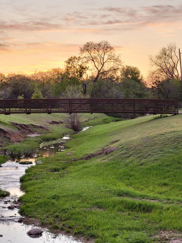 Photo of metal and wood pedestrian connecting green grassy banks of a stream in the foreground.  The background is treetops in the distance and a colorful evening sky, yellows, oranges, and pinks.