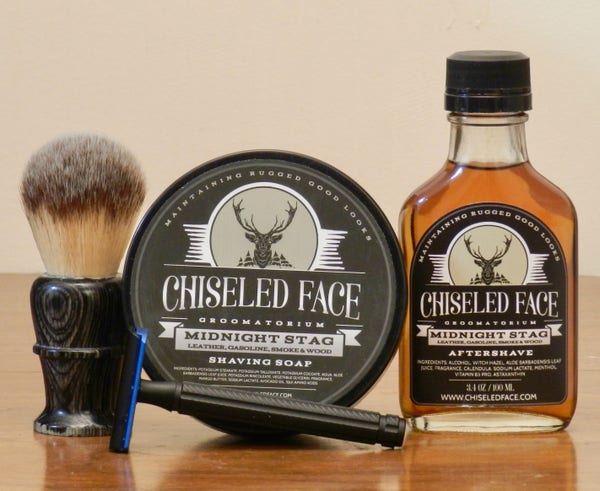 A shaving brush with a polo handle of treated wood dyed black and a light tan synthetic knot with dark-grey tips stands next to a tub of shaving soap whose black label has at the top a stylized drawing of the frontal vieeww of a stag's head with sillhouettes of evergreen trees in the distant background. Below that, in large white letters is "Chiseled Face." A representatin of a white ribbon is beneath, with "Midnight Stag" printed on it and, in smaller letters, "Leather, Gasoline, Smoke & Wood." Next to it is a clear-glass bottle with the same label and a black cap filled with an amber liquid.  Lying on its side in front is a black DE razor with a bright blue baseplate.