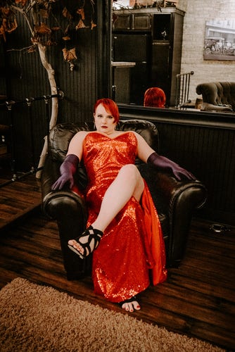 The poster, wearing a Jessica Rabbit dress, reclines n a plush brown leather chair with her legs crossed and looks directly at the camera. 