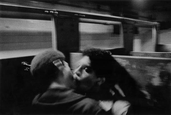 Black and white photo in a grainy style of two men kissing on a train
