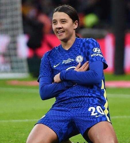 Sam Kerr in her blue Chelsea uniform, sitting I. The pitch with both knees on the ground, arms crossed and smiling