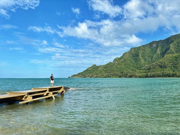 A person stands at the end of a wooden pier extending into a turquoise sea with mountains and a clear blue sky in the background.