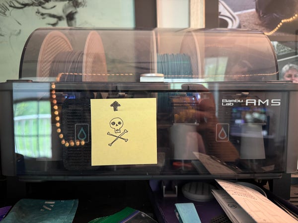 A photograph of a Bambu Lab AMS  (automatic, multi-filament selector)

A Post-it note with a skull and crossbones drawn on it beneath an arrow that points upwards to an empty slot in the AMS