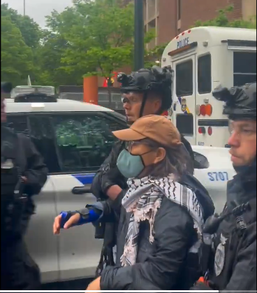 A student in an orange baseball cap, blue covid mask and wearing a keffiyeh is flanked by two police in tac gear.