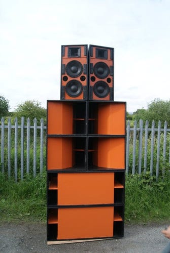 A stack of orange and black speakers in front of a metal fence