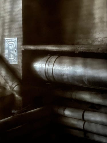 Colour photo, in muted tones of beige and grey, of some very-industrial-for-Venice shiny metal pipes affixed to the wall of a narrow alleyway. The photo is taken looking out of a window through mosquito netting, which gives a hazy noir-ish feeling to the image. 