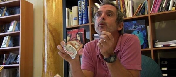 Photo de John B. Root, assis devant des étagères remplies de livres. Il porte une montre noire au poignet droit et une chemise rose. Les étagères en bois contiennent une variété de livres, donnant l’impression d’une bibliothèque ou d’un espace de lecture personnel.