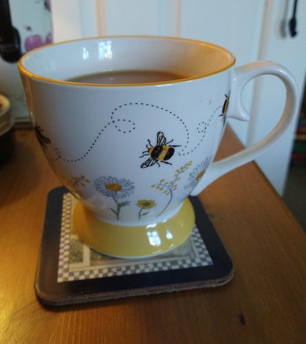 Pretty bowl shaped mug of tea on a bedside cabinet, it has a yellow base, at the bottom are daisy like flowers and bees above 
